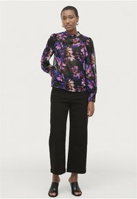 Transparente schwarze Bluse mit floralen Motiven in Lila und Pink, langen puffigen Ärmeln; kombiniert mit schwarzen weit geschnittenen Hosen und offenen schwarzen Mules.