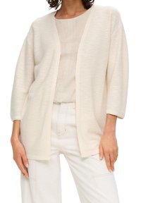 Světlý béžový otevřený kardigan z texturované látky s volnějším střihem, 3/4 rukávy a bez zapínání. Nošený přes lehké tričko a kalhoty.