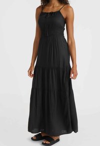 Kvinna i en ärmlös svart lager-på-lager maxiklänning med tunna axelband och svarta klippsandaler, stående framför en vit bakgrund.