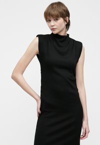 Jauna sieviete ar īsiem brūniem matiem, tērpusies melnā bezceļojuma augstās kakla kleitā, stāvot pretī gaiši pelēkam fons.