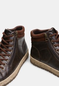 Brown leather high-top shoes with a zip feature, lace closure, and beige rubber sole, designed with suede accents and textured surface.
