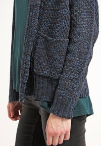 Tummansininen neulecardigan, jossa on ribbireunat hihoissa ja helmassa, teksturoidulla kuviolla ja yhdessä etutaskulla, kerroksittain vihreän paidan päällä.