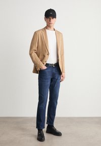 Blazer di lino beige sopra una t-shirt bianca, jeans blu scuro, cappellino nero e mocassini neri con dettagli dorati. Il modello sta in piedi con le mani nelle tasche.