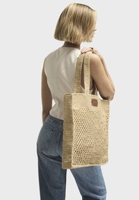 Borsa tote beige intrecciata con spalline spesse e un motivo testurizzato. Presenta un dettaglio in pelle. Modello che indossa jeans larghi e un top aderente.