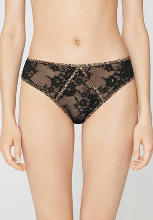 Femme portant une culotte en dentelle florale noire avec un bord imprimé léopard, montrée de la taille jusqu'aux cuisses sur un fond neutre.