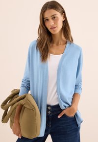 Vrouw met lang bruin haar in een lichtblauwe cardigan, witte top en donkere jeans, die een grote gewatteerde olijfgroene schoudertas vasthoudt.