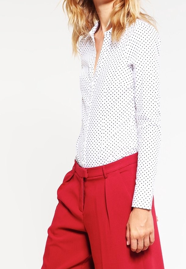 Femme portant une chemise blanche à manches longues avec des pois noirs et un pantalon rouge taille haute, main reposant à côté.