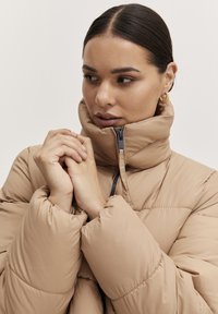 Tan puffjacka med hög krage, zippered front och pösig textur. Innehåller elastiska ärmslut och en subtil glans för ökad visuell intress.