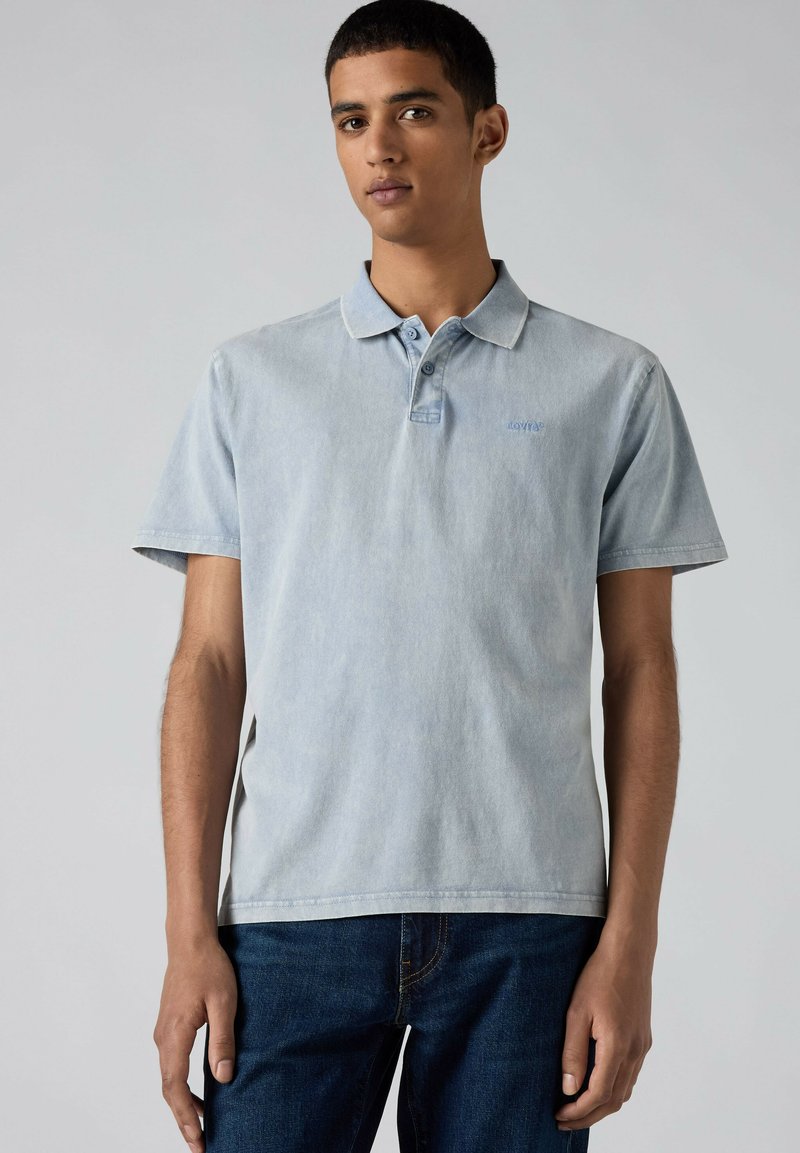Young man wearing a light blue short-sleeve polo shirt and dark blue jeans standing against a plain gray background.
