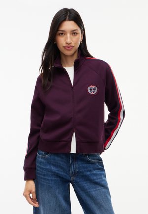 Kvinde med langt mørkt hår iført en lilla lynlåstrøje med røde, hvide og sorte striber på ærmerne og blå jeans mod en hvid baggrund.