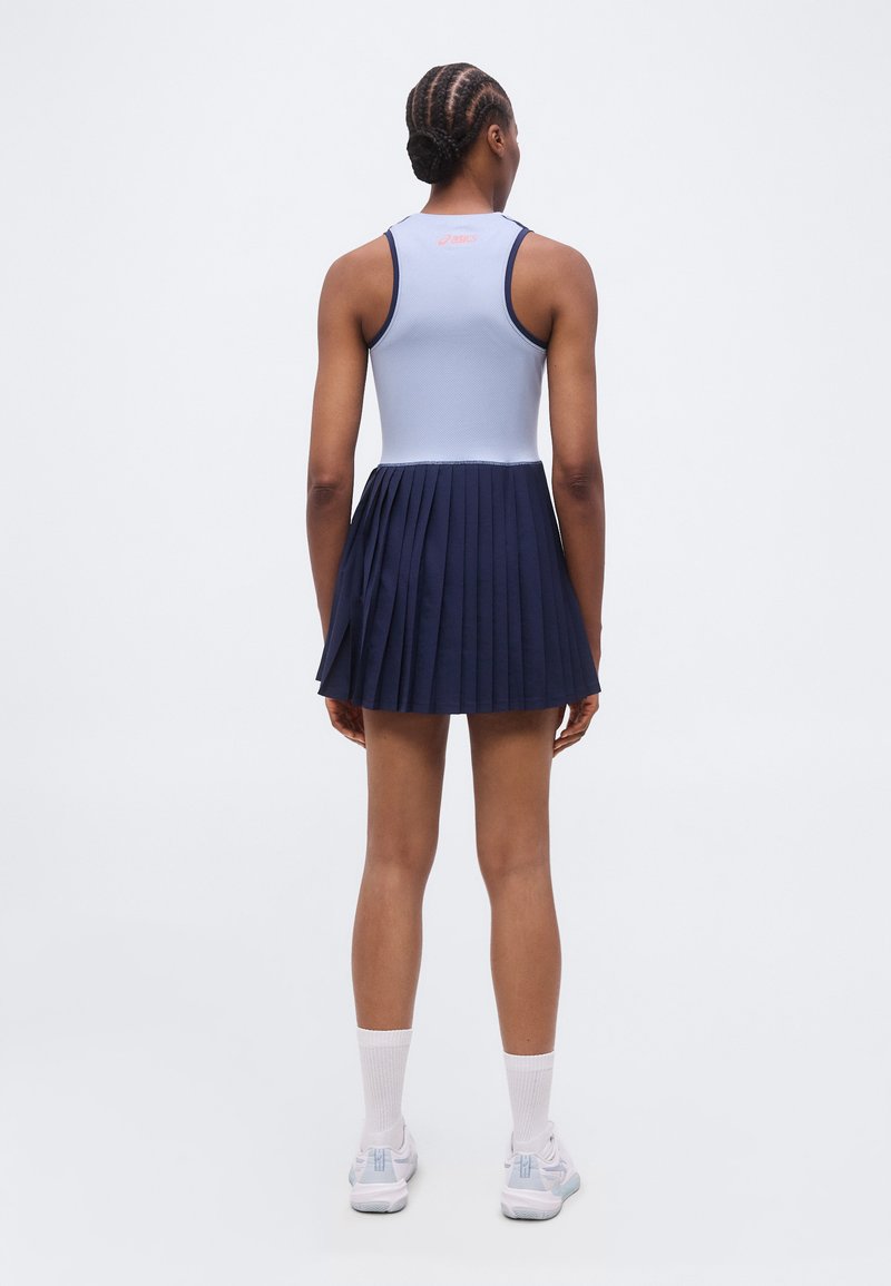 Frau mit geflochtenem Haar, die ein ärmelloses hellblaues Top und einen marineblauen plissierten Rock trägt, mit dem Rücken zur Kamera stehend.