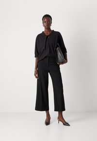Zwarte blouse met een striknek, losse mouwen en textuur. Gecombineerd met zwarte high-waisted broek met wijde pijpen en zwarte puntige hakken.
