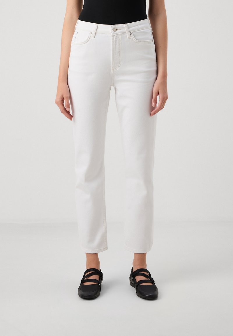 Vaqueros blancos de mezclilla con pierna recta, cintura alta y diseño de cinco bolsillos. Combinados con zapatos planos negros con tiras cruzadas.