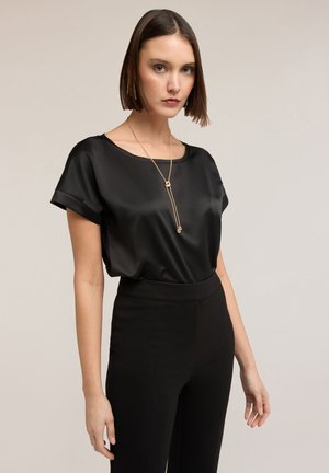 Kvinde iført en sort satin kortærmet bluse, højtaljede sorte bukser, guldkæde og lange guldbøjler mod en beige baggrund.