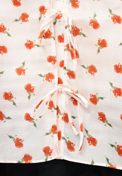 Φλοράλ μπλούζα με υφασμάτινη υφή σε λευκό χρώμα, διακοσμημένη με πορτοκαλί λουλούδια και πράσινα φύλλα, δεμένη μπροστά με κορδόνια από το ίδιο ύφασμα.