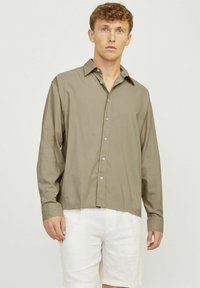 Camisa de manga larga verde oliva en tejido suave, con botones en la parte delantera y cuello puntiagudo. Combinada con pantalones cortos de color claro.