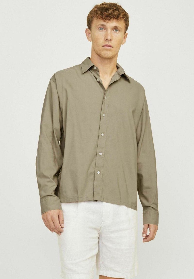 Camisa de manga larga verde oliva en tejido suave, con botones en la parte delantera y cuello puntiagudo. Combinada con pantalones cortos de color claro.