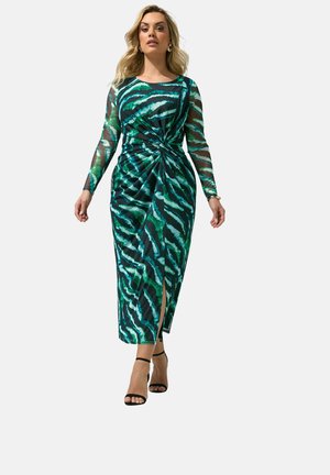 Vrouw draagt een lange mouwen, groen en zwart zebraprint jurkje met een zijsplit en zwarte enkellaars hakken, loopt zelfverzekerd vooruit.