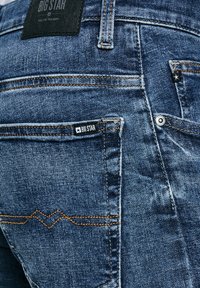 Blaue Denim-Jeans mit orangefarbener Naht, Big Star Markenlabel auf der Gesäßtasche und am Bund, Nahaufnahme zeigt Textur und Details.