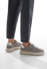 Baskets en daim gris avec un design texturé, accents blancs et une semelle chunky beige, associées à un pantalon foncé coupé.