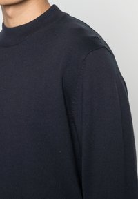 Maglione blu navy lavorato a maglia con scollo rotondo, caratterizzato da un tessuto strutturato e una scollatura a coste. Primo piano della spalla e del dettaglio della manica.