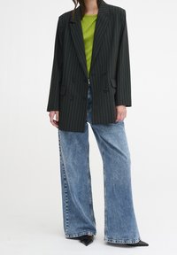 Czarny oversize żakiet w prążki z podwójnymi guzikami, zestawiony z jasną zieloną bluzką i spodniami dżinsowymi o szerokich nogawkach w kolorze niebieskim, uzupełniony czarnymi szpilkami.