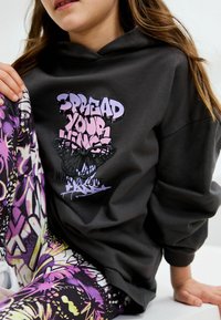Hoodie noir avec un texte graphique en violet, présentant un design de papillon. Associé à un legging à motifs vibrants dans des tons de violet et de jaune.