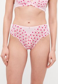 Lacey roze slipjes met hartpatronen, golvende randen en een decoratieve knoopdetail aan de voorkant. Zachte, doorzichtige textuur.