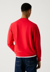 Mężczyzna ubrany w jednolitą czerwoną bluzę z długim rękawem i ciemnoniebieskie dżinsy, stoi tyłem na jednolitym jasnym tle.