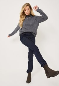 Pullover grigio a coste con collo alto, abbinato a jeans in denim scuro. Stivaletti marroni alla caviglia con tacco robusto completano l'outfit.