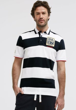 Mann trägt ein schwarz-weiß gestreiftes Poloshirt mit einem GA 97 Aufnäher auf der linken Brust, steht mit einer Hand in der Tasche.