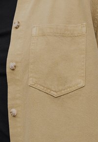 Μπεζ τζιν πουκάμισο μπροστά με ορθογώνια τσέπη στο στήθος και τρία ανοιχτόχρωμα κουμπιά στην αριστερή πλευρά πάνω από μαύρο ένδυμα.