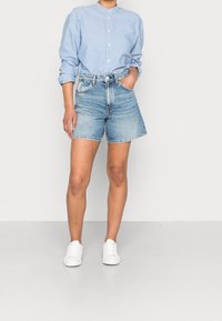 Person klädd i en ljusblå knappad skjorta med uppvikta ärmar, högmidjade denimshorts och vita sneakers som står framför en enfärgad bakgrund.