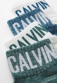 Ponožky Calvin Klein v troch farbách: bielej, tyrkysovej a námorníckej. Každé majú pletené biele logo „CALVIN“ a rôzne vzory. Mäkka textúra, rebrované manžety.