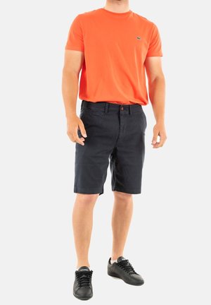 T-shirt arancione a girocollo, pantaloni corti navy con tasche frontali e chiusura con bottone, abbinati a sneaker nere con una texture liscia.