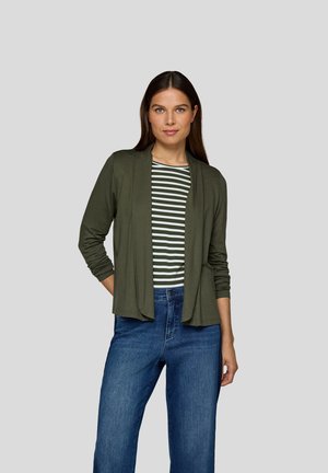 Femme avec de longs cheveux bruns portant un cardigan ouvert vert olive, une chemise rayée verte et blanche, et un jean bleu, se tenant devant un fond uni.