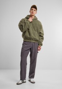 Olivgrüner Fleece-Hoodie mit Kängurutasche, kombiniert mit hellgrauen Cordhosen. Das Model trägt weiße Sneakers mit schwarzen Streifen.
