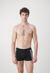 Junger Mann mit lockigem Haar und ohne Hemd steht frontal und trägt schwarze Boxershorts mit zwei weißen Seitenstreifen.