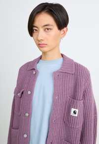 Lila gestrickte Strickjacke mit Knopfverschluss, umgeschlagenem Kragen und aufgesetzter Tasche. Getragen über einem hellblauen T-Shirt. Silberne Hardware-Akzente.