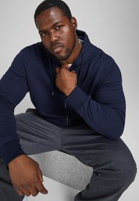 Marineblaues Zip-Hoodie mit Kordelzügen, kombiniert mit grauen gemusterten Hosen. Glatte Textur mit lässiger Passform.