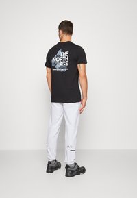 Svart t-shirt med vitt logotyp och bergsgrafik på ryggen, kombinerat med vita byxor och svarta skor, visar en avslappnad outfit.