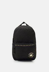 Sac à dos en tissu noir avec une poche frontale zippée ornée de clous dorés et d'un logo. Forme arrondie avec une poignée supérieure pour le transport.