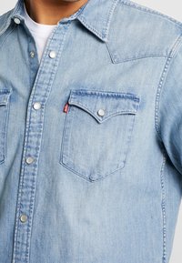 Närbild av en person som bär en ljusblå denimskjorta med tryckknappar och en bröstficka med en liten röd etikett.