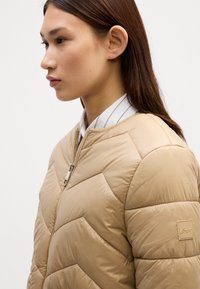 Beige pufferjakke med et chevronmønster. Har en lynlåslukning og et lille logo patch på ærmet. Glat, skinnende tekstur.