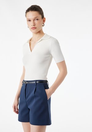 Donna che indossa un top bianco a maniche corte a coste con colletto e pantaloncini blu navy a vita alta con cintura, mano in tasca, su sfondo uniforme.