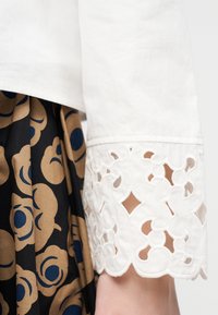 Λευκό μανίκι με περίτεχνη δαντελένια διακοσμητική λεπτομέρεια, σε συνδυασμό με μαύρο ύφασμα που φέρει μεγάλα καφέ και μπλε λουλουδένια μοτίβα.