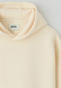 Sweat à capuche beige oversize avec une large capuche et une texture douce, présentant une étiquette marquée "BZB" à l'intérieur du col.