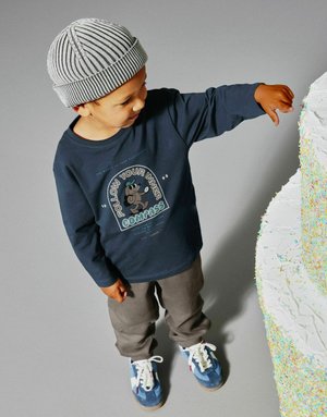 Młody chłopiec w paskowanej czapce i granatowej koszuli sięga w stronę dużego białego ciasta z kolorowymi posypkami na szarym tle.