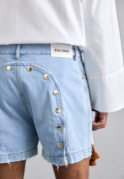 Shorts en denim bleu clair avec un ourlet effiloché, présentant des boutons dorés sur le côté, et une étiquette blanche avec "PALOMO" à la taille.