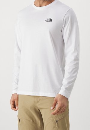 Mann trägt ein weißes Langarmshirt mit dem The North Face-Logo auf der Brust und beige Hosen mit Reißverschlusstaschen.